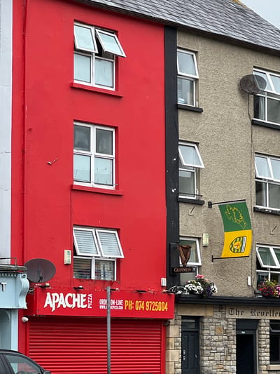 The image depicts the facade of two adjoining buildings in the town of Donegal, Ireland. The building on the left is painted a vibrant red and features several windows, some open and some closed. A red shuttered storefront with the "Apache Pizza" logo is visible at the bottom. The building on the right is a muted tan color with a rough stucco texture. It has multiple windows, some open, and a satellite dish mounted on the exterior wall. A prominent green and yellow flag, possibly representing a county or sports team, hangs from the wall. Below the flag, a Guinness sign is mounted. Flower boxes filled with colorful flowers are situated on the ledge between the two buildings. The upper story of the tan building shows a section of a grey, slate-tiled roof. The entrance to a pub or establishment named "The Reveller" is visible on the lower level of the tan building, constructed with stone blocks. The scene is captured on an overcast day, suggesting a cloudy sky.