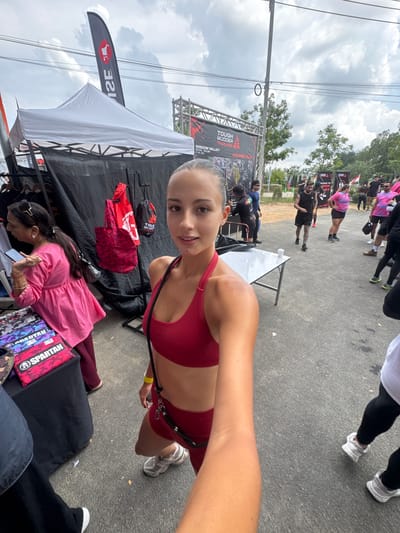 Here is a description of the image:
The image appears to be a selfie taken outdoors at an event, likely a race or competition, in บ้านสำนักตะแบก, Thailand. The person taking the selfie is a young woman wearing a red sports bra and shorts. She has fair skin and her hair is pulled back. She is smiling slightly at the camera.
In the background, there is a crowded area with many people walking around. There is a banner that says "TOUGH MUDDER THAILAND, PATTAYA, THAILAND, WISDOM VALLEY" indicating the event and location. There are also some tents and tables set up. One tent has a white canopy and a flag with a red logo.
Another woman is bending over a table in front of the tent, looking at something, possibly a product or item for sale. She is wearing a pink top and has her hair down. There are various items on the table. The ground is paved and there are trees and power lines visible in the distance.