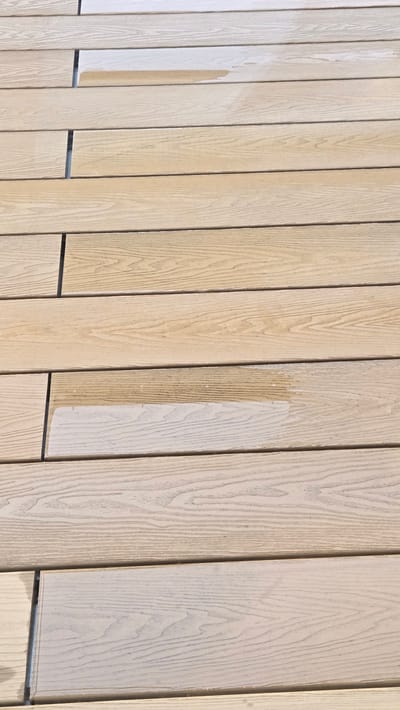 The image shows a close-up, high-angle view of a wooden deck with horizontal planks. The wood appears to be a light, natural tone with visible grain patterns. Some of the planks have a glossy, wet sheen, indicating the presence of liquid, likely water. The planks are evenly spaced, with thin, dark lines visible between them. The scene is devoid of people or any distinguishing features that would suggest a specific location within Gwangmyeong, South Korea. The focus is entirely on the texture and surface of the wooden deck.