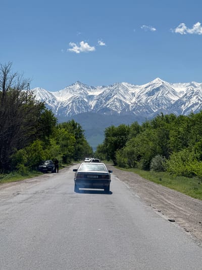 A photograph taken in Tokmok, Kyrgyzstan, shows a gray asphalt road running diagonally from the lower right to the upper right. On the left side of the road are green trees and shrubs, with a snow-capped mountain range visible in the background. A bright blue sky with a few small clouds is present. A silver car is traveling down the road away from the viewer. There is a dark colored vehicle stopped on the side of the road.
