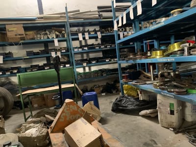 The image depicts the interior of a cluttered industrial or commercial storage facility located in Babawa, Nigeria. Multiple tall, blue, metal shelving units are visible, extending into the background, densely packed with various items. The shelves on the left predominantly hold small metal components, fittings, and cardboard boxes, while those on the right display a wider assortment of metal parts, including flanges, discs, and cylindrical components, some appearing brass-colored or worn. White labels are affixed to many shelves. The concrete floor is unkempt, with numerous objects scattered throughout the foreground and midground. These include a large vehicle tire on the left, a green metal workbench with a cardboard box on its surface, a tall black paddle-like tool, two blue plastic jerricans, an orange triangular object, several cardboard boxes (one prominently labeled "PHILIPS Stefan 867"), and a black plastic trash bag. Large white plastic containers are visible beneath the shelves on the right. The overall scene suggests an active but disorganized storage area for industrial parts and equipment. No individuals are present in the image.