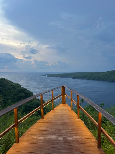 This image captures a wooden viewing platform situated on a forested clifftop overlooking a body of water, identified as a coastal area in Sakti, Indonesia. The platform, constructed with horizontal wooden planks, forms a triangular shape extending towards the viewer. It is bordered by wooden railings composed of dark vertical posts and lighter horizontal handrails, which feature integrated linear yellow lighting along their interior surfaces. No individuals are present on the platform. Below the platform and along the adjacent slopes, dense green foliage is visible. The midground consists of a wide expanse of dark blue, rippling water, consistent with an ocean or bay, bordered by lushly forested landmasses curving into the distance on both the left and right. A faint white structure is discernible on the distant left coastline. The background sky is overcast with predominantly blue and grey clouds, exhibiting a subtle orange-pink gradient near the horizon, indicating either dusk or dawn lighting.