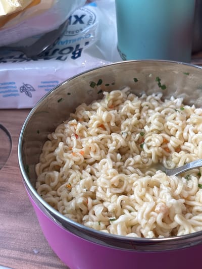The image captures a close-up, slightly elevated view of a meal in Tatarsk, Russia. The primary subject is a circular insulated bowl with a bright pink exterior and a silver metallic rim, occupying the lower and central portion of the frame. The bowl contains a mass of cooked, light-yellow instant noodles, distinguished by their wavy, helical structure. Scattered throughout the noodles are small green fragments, consistent with chopped chives or similar herbs, and occasional minute orange fragments, likely diced vegetables. A silver spoon is partially submerged within the noodles on the right side of the bowl, indicating active consumption or preparation.
In the blurred background, to the upper left, a white plastic bag featuring Cyrillic text, including the word 'ВОДА' (water) and a large 'X', is visible. Beneath this bag, a smaller white package, potentially containing napkins or wipes, also displays Cyrillic text and an illustrative leaf pattern. To the upper right, a portion of a light blue or teal insulated cup or bottle is present. The objects rest upon a light brown surface, consistent with a wooden table or countertop. No individuals are visible in the scene.