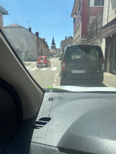 This is a photograph taken from inside a vehicle, looking forward through the windshield at a street scene. The windshield has a textured, dirty appearance. The street is lined with buildings on either side, mostly two to three stories tall, with some architectural details suggesting a European setting. The sky is clear and blue.
In the foreground, the dashboard of the car is visible, with a dark gray, textured surface. There is an air vent on the left side. On the dashboard, near the windshield, there are some green stickers with text, and a black circular emblem.
On the street, there are two cars. The closest one is a dark-colored van, possibly black or dark gray, with its rear facing the camera. The license plate of the van is visible and reads "DC 761 00". Behind the van, further down the street, is a small, bright red car, also facing away from the camera. The red car appears to be a classic model.
There are white pedestrian crossing lines on the asphalt road. In the distance, a church steeple with a dark roof is visible between the buildings. The overall impression is of a sunny day in a small town or village. The presence of the "DC" prefix on the license plate indicates it is a French license plate. Given the architecture and the style of the cars, this location is likely in France. Specifically, the license plate format suggests it could be from a department that historically used "DC" as a prefix, such as the Haute-Saône or the Jura department. A reverse image search or knowledge of French license plate evolution might pinpoint the exact location, but the prompt states "Coteaux du Lizon, France" as the location. Coteaux du Lizon is a commune in the Jura department in Bourgogne-Franche-Comté in eastern France. This is consistent with the "DC" license plate.