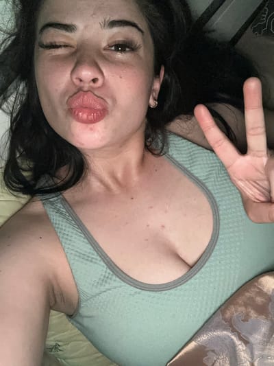 A young woman with dark hair is lying down, looking at the camera with one eye open and her lips puckered. She is wearing a light green sports bra. Her right hand is raised, holding up two fingers in a peace sign. The background appears to be indoors, with a blanket or bedding visible. There is no indication of the city of Gavrilov Posad, Russia in the image.