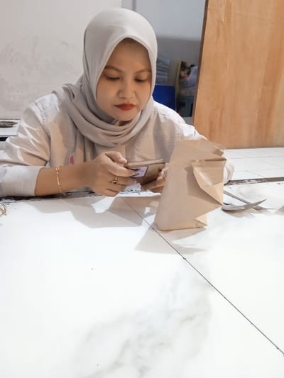 An indoor, eye-level shot captures a woman sitting at a light-colored table in Cangkring, Indonesia. She is wearing a light gray hijab and a matching long-sleeved shirt. Her face is directed downwards, focused on a smartphone she holds with both hands. She has red lipstick. On her left wrist, she wears a gold-colored bracelet, and a ring is visible on her right hand.
To her right, on the table, stands an upright brown paper bag, commonly used for food packaging. Beside the bag, a silver-colored fork and spoon are placed on the table. The table surface is bright white, possibly tiled, with a dark line running diagonally across the lower right section. The background features a plain, light-colored wall on the left and a light brown wooden panel or door on the right. Some indistinct items are partially visible in the middle background. The lighting is bright, suggesting an indoor setting during daytime or well-lit conditions. The woman appears calm and engaged with her phone, possibly awaiting or pausing a meal.