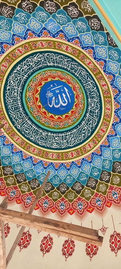 This image depicts a large, intricately painted circular Islamic mural located on the interior ceiling or dome of a structure in Kumbang, Indonesia. The central element is a blue disc featuring the Arabic word "Allah" (الله) in white Thuluth script, with a smaller Arabic word beneath it. This central disc is surrounded by multiple concentric rings, each adorned with distinct patterns and Arabic calligraphy.

Immediately around the central blue disc is a red ring with small, light-colored floral or geometric motifs. This is followed by a teal ring containing continuous white Arabic calligraphy. Subsequent rings display a light green band with red and blue patterns, a dark blue band with lighter blue and white stylized motifs, and then rings of teal and olive-brown backgrounds, both segmented with white Arabic calligraphy in stylized, often leaf-like shapes. The overall color palette is vibrant, featuring blues, greens, reds, gold, and white.

The mural is set against a light-colored, possibly off-white, ceiling. In the lower left and central portions of the image, two rough-hewn wooden beams are visible, angled diagonally upwards from left to right, partially obscuring the lower section of the artwork. A thin, silver-colored metal chain hangs vertically from the central blue disc, passing over the white "Allah" calligraphy. A light blue architectural element is visible along the upper right edge of the frame. No individuals are present in the scene.