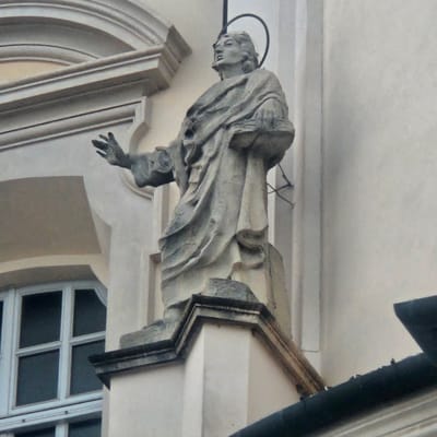 The image shows a low-angle, medium shot of a weathered stone statue of a saint, identified as Saint Victor, located on the exterior of the Saint Victor and Saint Corona Church in Calcinate, Italy. The statue depicts the saint with a halo, draped in a robe, and holding a book. His left hand is outstretched, and his head is tilted upwards. The statue is mounted on a decorative pedestal against the pale facade of the church. A portion of a window with multiple panes is visible to the left of the statue, and a dark roofline runs along the bottom right of the frame. The lighting suggests it is daytime, with diffused light casting soft shadows.