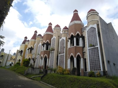 The image presents a residential street scene in Nathene, Laos, featuring a row of multi-story buildings with a distinct, elaborate architectural style. These structures are situated on a grassy slope ascending from a paved, dark gray road in the foreground. Each building unit displays facades in varying shades of cream, beige, and brown, accented by multiple white columns, arched windows and doorways, and prominent patterned decorative screens on vertical panels. Most units feature red, conical or onion-shaped cupolas atop their structures, with some incorporating upper-story recessed balconies framed by arches and columns. The building at the far right exhibits a plain, unadorned gray side wall. The ground cover consists of green grass interspersed with sparse shrubbery. The sky above is partly cloudy with visible blue patches. No individuals are present in the frame.