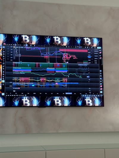 The image shows a close-up, eye-level, medium shot of a television screen displaying a cryptocurrency trading chart, specifically for Bitcoin (BTC) against the US Dollar. The chart features multiple indicator lines in various colors, including purple, pink, blue, and green, overlaid on a black background. The screen is positioned in front of a wall with a textured, light-colored finish that appears to be a light beige or pale pink marble effect. Below the television screen, a portion of a light-colored, modern furniture piece is visible, possibly a cabinet or credenza. The overall setting appears to be an indoor environment, likely a home or office, with ambient lighting. The television screen is emitting light, illuminating the immediate surroundings. There are no visible people, and the focus is entirely on the digital display of financial data. The image is taken from a position that suggests the viewer is standing or sitting directly in front of the television. The specific location of Oberleichtersbach, Germany, is not discernible from the visual information presented in the image.