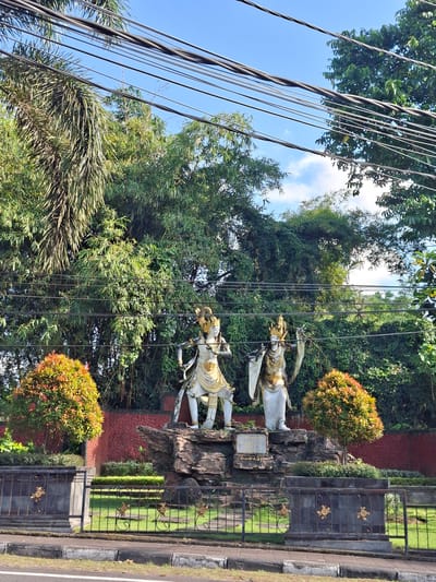The image depicts an outdoor scene in Banjarangkan, Indonesia, with a prominent display of two ornate statues situated on a rocky base. These statues, appearing to be made of stone or a similar material, are adorned with gold accents and elaborate headdresses, each holding a distinct object or pose. One statue stands with a sword-like object, while the other has flowing drapery. Behind the statues, a lush green bamboo forest forms a dense backdrop, interspersed with other foliage. Several thick electrical wires are strung across the upper portion of the frame, silhouetted against a bright blue sky with scattered white clouds. In the foreground, a black metal fence with decorative gold floral motifs encloses a manicured green lawn. Two spherical, vibrantly colored bushes, one orange-red and the other with mixed green and red leaves, flank the statue display on either side. A section of a red textured wall is visible behind the bushes and to the left of the statues. The lower edge of the image shows a paved road and sidewalk, suggesting a location along a street.