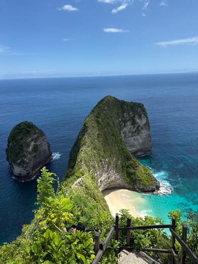 This image captures a scenic view of Kelingking Beach, located in Bunga Mekar, Indonesia. The dominant feature is a large, T-shaped land formation covered in lush green vegetation. This formation extends into the deep blue ocean, creating a sheltered cove with a sandy beach. To the right of the frame, a wooden fence and dense green foliage suggest a viewing point on a cliff. Below the main land formation, a smaller rocky island is visible in the ocean. The sky is clear blue with a few scattered clouds. Waves are crashing against the shore.