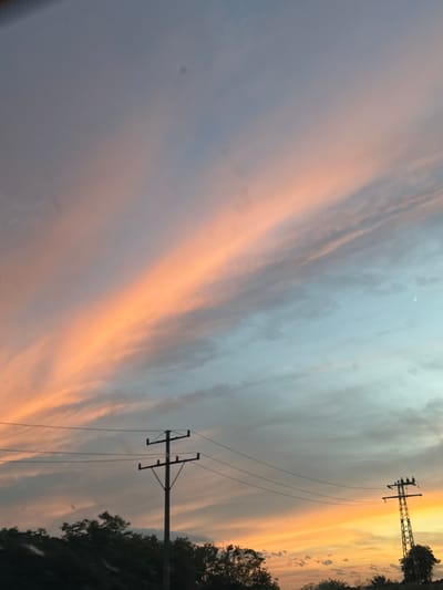 The image depicts a sky with wispy clouds tinged with orange and pink hues, characteristic of a sunset. The sky transitions from a soft gray at the top to a pale blue and then to a vibrant orange near the horizon. Silhouetted against the sky are trees, power lines, and two utility poles. The foreground shows the dark shapes of trees and foliage. In the upper right portion of the sky, a faint crescent moon is visible. The scene appears to be set in an outdoor environment, possibly a roadside or open area, with signs of infrastructure like power lines. While the image was captured in the city of Tvardintsi, Bulgaria, there are no specific landmarks visible that would definitively identify the exact location within the city.