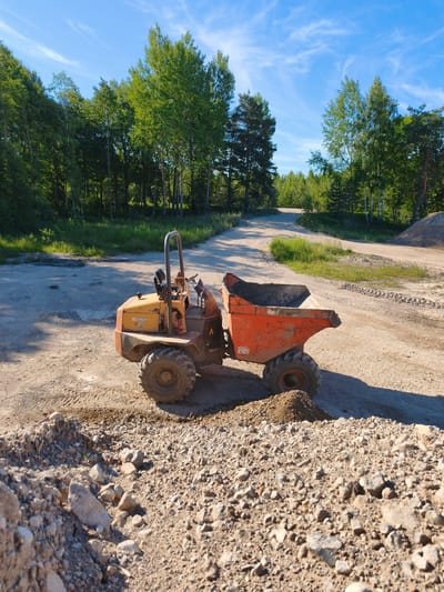 An orange and yellow dumper truck sits on a dirt road surrounded by trees and blue sky with white clouds. The truck is in the foreground, with its front facing left and its bed angled towards the right. The bed is empty. The dumper truck appears to be on a construction site or a rural road, with piles of dirt and gravel visible. The scene is lit by sunlight, creating shadows on the ground. The location is Skrunda-1, Latvia.
