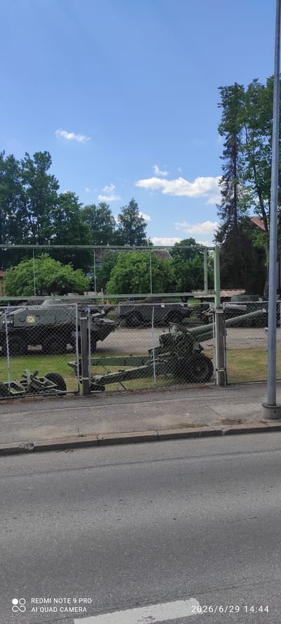 In Valga, Estonia, a clear blue sky with scattered clouds is visible above a fenced area containing military vehicles. The fence, made of chain-link, runs horizontally across the image, separating the viewer from the display. Inside the enclosure, several military vehicles, including what appear to be tanks and artillery pieces, are parked on a grassy area. Trees form a backdrop behind the vehicles, with some buildings also visible through the foliage. In the foreground, a paved road and sidewalk are present, indicating this scene is likely accessible from a public area. The overall impression is that of an outdoor military museum or exhibition.