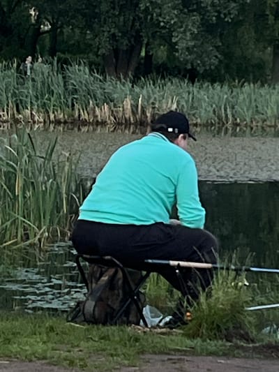 A person is fishing by a body of water, surrounded by tall grass and trees. The person is wearing a turquoise long-sleeved shirt and black pants, and a black baseball cap. They are sitting on a black folding stool with a camouflage bag underneath. Two fishing rods are placed in front of them, extending towards the water. The water is dark and reflects the greenery of the surroundings. In the background, more reeds and trees are visible, with two distant figures walking near the trees. The ground in the foreground is covered in green grass and some brown dirt.