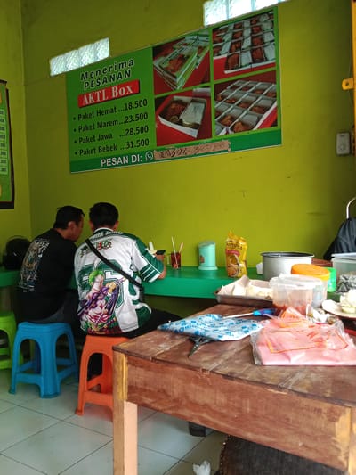 Two men are seated at a green table within a small, casual eatery in Sekopek Wetan, Indonesia. One man wears a black t-shirt with a graphic on the back, and sits on a blue plastic stool. The other man is wearing a white and green shirt with a "One Piece" themed design and a shoulder bag, seated on an orange plastic stool. The eatery has green walls, and a poster displaying "AKTL Box" meal deals is visible, along with a phone number for orders. Various condiments, a bag of cooking oil, and other food-related items are arranged on the green table. In the foreground, a large, worn wooden table holds items like plastic bags, scissors, and food containers. The environment suggests a local, inexpensive food establishment.