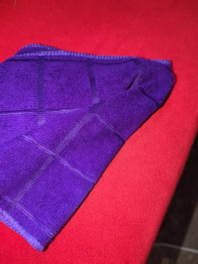 A close-up, high-angle shot shows a folded purple microfiber cloth with a grid pattern laid diagonally on a bright red textured surface. The cloth's edge, with its reinforced stitching, is visible along the top left of the frame. The red surface appears to be fabric, possibly felt or a similar material, with a fine, uniform texture. The lighting creates a slight shadow along the bottom right edge of the frame, suggesting the light source is from the top left. The scene is devoid of people or any indication of human activity. There are no identifiable landmarks or elements that would suggest a specific location within Elele Alimini, Nigeria.