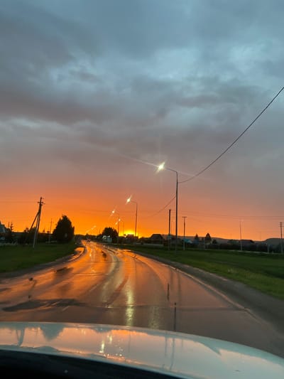 The image depicts a wet, paved road curving through a rural landscape during a vibrant sunset. The sky is a dramatic gradient of deep gray clouds overhead, transitioning to a brilliant, fiery orange closer to the horizon, with the sun itself not directly visible but implied by the intense illumination. The road surface reflects the intense orange of the sky, creating shimmering patterns. Streetlights, illuminated and reflecting on the wet asphalt, line the right side of the road, spaced at regular intervals.
To the left and right of the road, there are grassy verges and a few trees, some of which are dark silhouettes against the bright sky. In the distance, along the sides of the road, are glimpses of houses with pitched roofs, suggesting a small settlement or village. A power line with insulators runs diagonally across the upper right portion of the image, and another utility pole with wires is visible on the left. A white vehicle's hood is prominent in the foreground, partially obscuring the road and reflecting the sunset colors. The overall impression is that of a journey through a scenic, post-rain environment at dusk.
The location is stated to be in the city of Субханкулово, Russia.