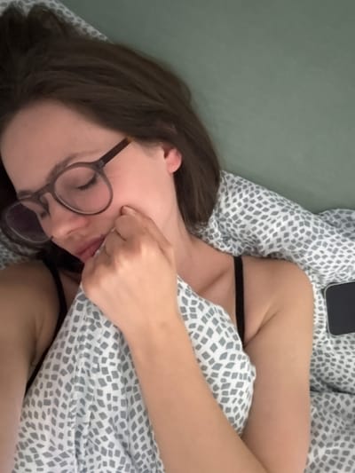 A woman with brown hair and wearing glasses lies in bed with her eyes closed. Her left hand is touching her face. A smartphone rests on the pillow above her head. The bedding has a light gray geometric pattern. The wall behind the bed is green. The location and city of Kranj, Slovenia cannot be confirmed in the provided picture.