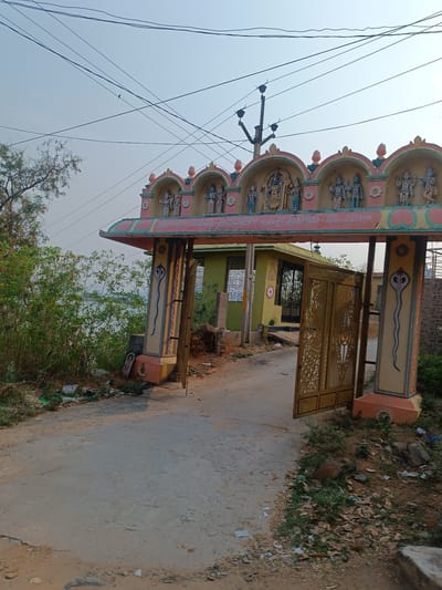 The image depicts the entrance to a temple in Kothacheruvu, India. The gate is ornate, with colorful carvings of deities along the top and snake motifs painted on the supporting pillars. The gate is partially open, revealing a small green building inside the temple grounds. The foreground shows a paved road leading to the gate, with scattered trash and foliage on either side. A telephone pole with electrical wires is visible above the gate. The sky is overcast. There are no people visible in the frame.