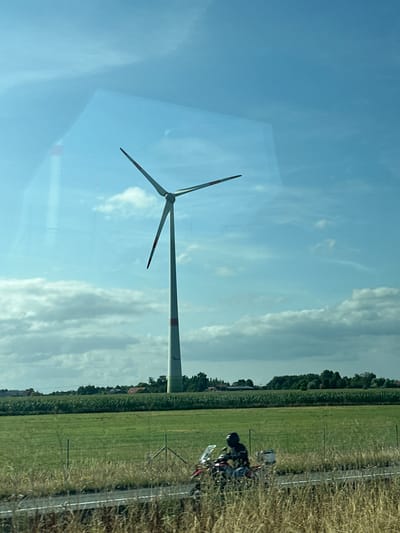 A vertical, slightly angled shot captures a tall wind turbine with three blades rotating against a blue sky dotted with scattered white clouds. The turbine is white with red bands near the top and bottom of its shaft, and some faint text is visible on the lower part of the shaft. In the foreground, a green grassy field stretches towards a line of trees and buildings in the distance. A motorcycle with a rider wearing a black helmet is visible on a road running through the field. The image is taken through a reflective surface, possibly a window, as indicated by faint reflections in the sky. The scene appears to be a rural landscape. The location is identified as Wuustwezel, Belgium.