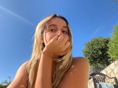 A medium close-up, low-angle photograph depicts a young female outdoors in Oliveira de Frades, Portugal, under a clear, bright blue sky with direct sunlight. The subject, positioned slightly off-center to the left, has long, light blonde hair and is looking directly at the camera. Her right hand is raised, with fingers covering her mouth and lower nose. Her right shoulder and arm are visible, exposed to the sun. In the background, green vegetation includes a prominent rounded tree with olive-green leaves and dense, dark green foliage to the right. A stone retaining wall constructed of irregular grey stones is visible behind the foliage. In the lower right quadrant, a dark brown or black plastic garden chair with a lattice-patterned back is partially visible, with a light blue towel draped over its seat. A faint aircraft contrail is present in the upper left portion of the sky, intersecting a sun flare. Utility lines traverse the upper right quadrant of the frame, above the trees. Strong shadows are cast on the subject, indicating direct overhead sunlight.