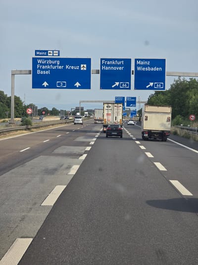 An overhead view captures a highway scene with multiple lanes of traffic under a hazy sky. Prominent blue highway signs provide directional information. One large sign indicates "Würzburg," "Frankfurter Kreuz," and "Basel," with arrows pointing straight ahead and a number "3" enclosed in a white rectangle. To the right, two other signs display "Frankfurt Hannover" and "Mainz Wiesbaden," each with an arrow indicating a turn and the number "66" in a white rectangle. Several vehicles, including cars and trucks, are visible on the highway. The road surface shows signs of repair with patched areas. The environment is urban with trees and vegetation visible along the roadside and in the distance, suggesting the location is near Breckenheim, Germany, given the highway signs.