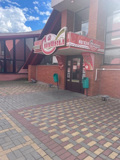 The image shows the exterior of a store with a red brick facade, located in Бабовіцкі сельскі Савет, Belarus. A large red and white sign above the entrance reads "МАГАЗИН СЛАДОСТИ" which translates to "SWEET SHOP". The store has a glass-paneled entrance door. To the left of the entrance, a red tiled roof structure with large windows is visible. The ground in front of the store is paved with interlocking bricks in shades of red, yellow, and grey. Two green trash cans are situated near the entrance. In the background, the sky is blue with scattered white clouds. The scene appears to be an outdoor shot on a sunny day.