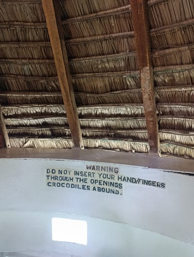 The image shows a close-up view of a warning sign attached to the underside of a thatched roof. The sign reads, "WARNING, DO NOT INSERT YOUR HAND/FINGERS THROUGH THE OPENINGS, CROCODILES ABOUND". The roof is made of woven palm leaves and supported by wooden beams. The sign is located in Tsavo North National Park, Kenya, near the Mzima Springs (latitude: -2.986, longitude: 38.023).  The scene appears to be inside a structure, likely a viewing platform or shelter, offering a vantage point to observe the Mzima Springs, a well-known spot where crocodiles are commonly sighted. The sign highlights the potential danger to visitors and provides a clear warning to avoid putting hands or fingers through any gaps in the roof.