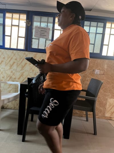 The image depicts an indoor or semi-enclosed commercial space in Abuja, Federal Capital Territory, Nigeria. A single individual, appearing to be female, is captured in profile, facing towards the right side of the frame. She wears a black baseball cap, an orange short-sleeved t-shirt, and black shorts with the word "Soccer" printed vertically in white cursive text on the left leg. Her right hand holds a black smartphone, while her left hand holds a crumpled black plastic bag. Both items are held together near her torso as she stands or moves.

The background features a wall with light brown, horizontally-banded tiling on the lower half and a lighter, possibly cream-colored painted surface above. Multiple windows with blue frames are visible, featuring internal grid patterns and allowing natural light to enter. A white paper sign is affixed to one window, displaying text including "SUI-GENERIS," "ACCT NO: 9650215827," "PALMPAY," and a phone number "08035464645."

Furniture visible includes a white plastic chair with a woven texture on the left, and a dark, possibly black or dark brown, woven-textured table in the center, partially obscuring another similar dark chair behind it. A standalone dark, woven-textured chair is positioned against the wall on the right. A white electrical outlet is mounted on the wall to the right. The floor is covered with light-colored tiles.
