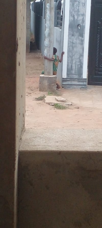 The image shows a young person in a green and orange patterned shirt lying on a concrete ledge, seemingly peering down. The location appears to be outdoors in Agunmo, Nigeria, given the context. The background includes a building facade and a dirt or concrete ground surface with sparse vegetation. A large concrete structure is visible to the right, partially obscuring the scene.