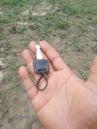 A hand, possibly belonging to a male, is holding a single car key with a black plastic head and a silver metal blade. The key is attached to a thin, black, braided loop. The hand and key are in the foreground, slightly out of focus. The background consists of dry, cracked earth with sparse green vegetation, suggesting a rural or arid environment. The text "Samsung Galaxy A03" is visible in the lower left corner of the image, indicating the device used to capture the photo. There are no other people or objects in the scene. The image depicts a common object in an outdoor setting. The location is the city of Angwan Hausawa, Nigeria.