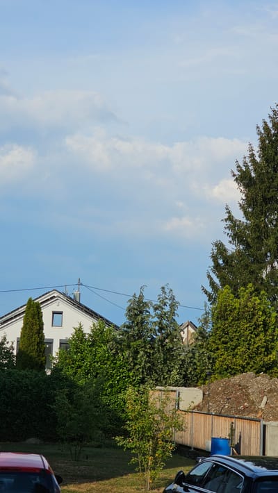 The image depicts an outdoor scene with a bright blue sky with scattered clouds, suggesting a sunny day. In the foreground, the top of a white house with a dark-framed window is visible. A conical evergreen tree stands in front of the house, and a small deciduous tree with green leaves is to its right. Behind the house, a wooden fence encloses a yard. A large blue barrel is positioned next to the fence, and a pile of debris, possibly construction material, is located further back. A portion of a dark-colored car is visible on the right side of the frame, and the rear of a red car is partially visible at the bottom right. Trees with dense green foliage frame the upper parts of the image. The scene is likely a residential area, possibly in Vöhringen, Germany, during daylight hours. There are no visible people or text in the image.