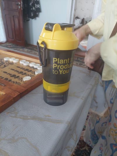 The image shows a yellow and black shaker bottle with the text "Plant to Produce to You" printed in yellow on its side. The bottle is placed on a table covered with a patterned tablecloth that has some light yellow stains on it. To the left of the bottle, a portion of a backgammon board with white checkers is visible. In the background, a dark wooden door with a metal handle and a dark garment hanging on it are visible. A person wearing a yellow long-sleeved shirt is partially visible on the right side of the image, with their hands near the shaker bottle. The environment appears to be indoors, possibly a home setting. The image does not contain any information about the city of Gusar, Azerbaijan.