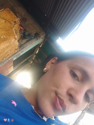 The image is a close-up, tilted, selfie-style shot of a young woman with dark hair. She is wearing a bright blue shirt with a pink ribbon-like design and what appears to be a clover-shaped pendant necklace. Her left ear is visible and adorned with a small, circular earring. The background suggests an outdoor, possibly rural setting in El Cercado, Venezuela, with corrugated metal roofing and rough, textured walls visible. A bright light source, possibly the sun, is creating a strong glare in the upper right portion of the image, obscuring some details. The woman's facial expression is slightly ambiguous, with her mouth partially open. The overall impression is of an informal, casual moment captured outdoors.