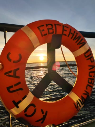 A close-up shot features a bright orange life preserver with white reflective stripes and Cyrillic lettering, prominently displayed against a backdrop of water and a setting sun. The text on the life preserver reads "ЕВГЕНИЙ ДРАБСКИЙ КОТЛАС," indicating the name of a ship or location. The life preserver is attached to a dark-colored metal railing and a support structure. The sun is setting on the horizon, reflecting light on the water's surface. The environment suggests a maritime setting. Considering the lettering on the life preserver and the general landscape, the location is likely Myaksa, Russia, a settlement near Kotlas, along the Northern Dvina River.
