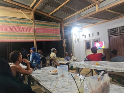 A group of men are gathered at tables, seemingly watching a television in an open-air structure with a corrugated metal roof supported by wooden beams. The structure appears to be informal, possibly a roadside eatery or community gathering space. One table in the foreground is covered with a marble-patterned cloth and holds plates of food, drinks, and a clear plastic container. Several men are seated at this table, one smoking. Further back, other men are seated at similar tables. The television in the background displays a person's face, possibly a sports player. The environment is dimly lit, suggesting it is nighttime or late evening. The location is identified as Rantopanjang, Indonesia.
