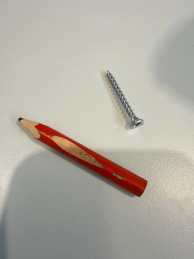 The image shows a red pencil with a sharpened, exposed graphite tip and a metallic screw with visible threading. The pencil is positioned diagonally from the bottom left to the upper right of the frame, with its sharpened end pointing towards the left. The screw is located in the upper right quadrant of the image, also oriented diagonally. Both objects are resting on a light-colored, textured surface, likely a desk or table. There are no people or any indications of a specific location in the city of Fontevivo, Italy. The lighting suggests an indoor environment.