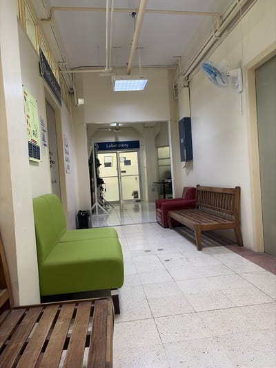 The image shows a hallway with a tiled floor. On the left, there is a bright green sofa and a sign that reads "Medical Records". Further down the hall, there is a sign that says "Laboratory" above a set of double doors. To the right of the hallway, there is a wooden bench and a red chair. A wall-mounted fan is visible on the right wall. The environment appears to be an indoor facility, possibly a hospital or clinic, given the presence of "Medical Records" and "Laboratory" signs. The location is not specified as Dagupan, Philippines.