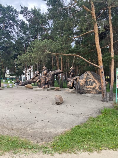 In Sosnovka, Russia, a park setting features a concrete paved area with a line of white planters and tall evergreen trees forming a backdrop. The ground is littered with small pinecones. In the foreground, a large sculpture depicts several bears. One prominent bear is seated on a concrete base next to a thick, dark-colored log that is angled upwards, supported by a concrete structure. Two smaller bears are perched on the log. Further to the right, another sculpture shows a bear standing on its hind legs, holding onto a thick rope that hangs from a tree branch. This bear is positioned next to a large, dark, irregular-shaped rock that rests on a concrete base. The overall scene is one of a decorative park installation in a natural environment.