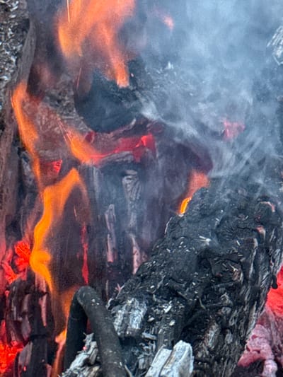 The image shows a close-up view of burning logs and smoke, with a prominent orange flame licking up the side of a dark, charred log. Ash and embers are visible throughout the scene, and wisps of grey smoke drift across the frame. The background is blurred, but appears to be a dark, textured surface, possibly more burning material or the ground. The scene conveys a sense of intense heat and combustion. The location in Imelevo, Russia, is not discernible from the image itself.