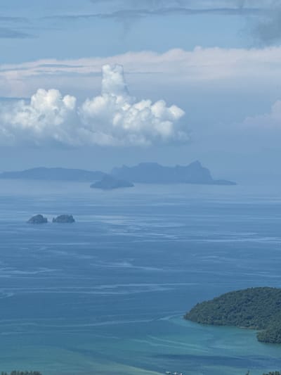 The image depicts a serene seascape with multiple islands in the distance. In the foreground, a lush green island juts into the turquoise water, creating a bay. The water exhibits subtle ripples and patterns. The middle ground features two small, rocky islands. Further in the distance, several larger islands are silhouetted against the hazy horizon. The sky above is partly cloudy, with fluffy white clouds against a pale blue backdrop. The overall impression is one of natural beauty and tranquility. The location is identified as Ban Din Daeng Nui, Thailand. There are no people or visible actions depicted in the image.