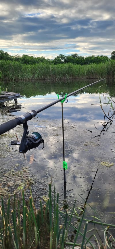 An outdoor, full-shot, eye-level photograph shows two fishing rods propped up to fish in a narrow canal with a backdrop of tall, green reeds and trees under a cloudy sky. The fishing rods are angled to the right of the frame. The rod in the foreground is black with a silver reel and is angled upwards and to the right, resting on its handle. The second fishing rod, positioned further back and to the right, is also black and is held up by a dark-colored rod rest with two bright green clips. The canal water is dark and murky, covered in patches of green algae and debris, with reflections of the sky and trees visible on the surface. Several stalks of green reeds are growing in the foreground and in the background, forming a dense wall along the canal's edge. To the far left, a portion of a wooden dock or pier is visible. The sky is filled with dramatic clouds, ranging from dark grays to lighter blues and whites. The location is identified as Hetmanivka, Ukraine.