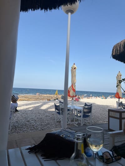 This is a vertical, eye-level shot of a beachside scene in Xàbia, Spain. The image depicts a relaxed atmosphere, likely during a summer day, with clear blue skies and calm ocean waters.
The foreground is dominated by the edge of a white structure, possibly a table or railing, and a black bag with circular cutouts and fringe detail. To the right, a wine glass with a small amount of white wine is prominent, alongside a bottle and a pair of sunglasses.
In the midground, several beach umbrellas are visible, some closed and tied, with cream-colored fabric and orange trim. Chairs with blue seats and white frames are arranged on a wooden deck. The beach itself is covered in white pebbles, and a few people are scattered along the shoreline and in the water. A yellow umbrella stands out on the beach.
The background shows the sea meeting the horizon under a bright, clear sky. The time of day appears to be late afternoon or early evening, given the angle of the sunlight casting shadows on the deck. The overall mood is one of leisure and vacation, typical of a coastal resort.
No visible text is present in the image.