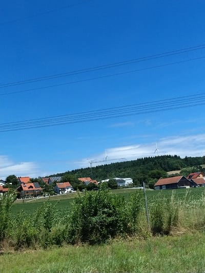The image depicts a rural landscape under a clear blue sky. In the foreground, tall green grass and shrubs line a pathway, with a silver pole visible on the right. Beyond the immediate foreground, a vast green field stretches towards a cluster of buildings in the mid-ground. These buildings are a mix of residential houses with red-tiled roofs and some modern structures, possibly with solar panels. Further in the background, a dense, dark green forest covers a rolling hill. Two white wind turbines stand prominently on the hilltop, indicating a nearby wind farm. The scene is likely located in or around Wörth an der Donau, Germany, given the architectural style of the houses and the presence of wind turbines, which are common in the German countryside. There are no people visible in the image, and the overall atmosphere is serene and natural.