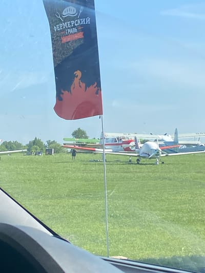 The image shows several light aircraft parked on a grassy field. A banner with Russian text and a fire image is visible in the foreground. The location is likely Chemodanovka, Russia. Details beyond these core observations are indiscernible.