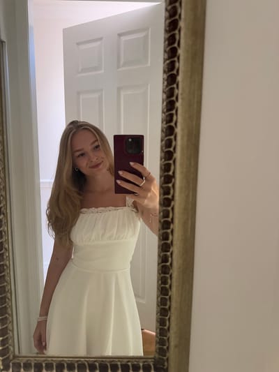 A single female subject, appearing to be a young adult with long, light brown hair, is depicted in a mirror selfie. She is wearing a white, strapless or spaghetti-strap dress with a ruched, lace-trimmed bodice and a flowing skirt. Her left hand, adorned with multiple rings, holds a dark-colored smartphone, consistent with an iPhone model, positioned to capture her reflection. A multi-strand bracelet is visible on her right wrist. The mirror features an ornate, textured frame. Reflected in the background is a white paneled door with three visible rectangular panels. This scene is located in Area B (Shawnigan Lake), Canada.