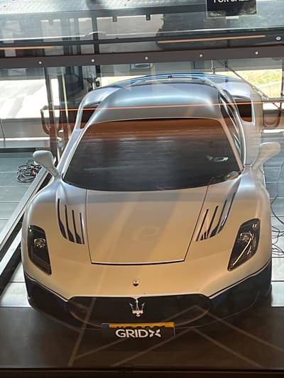 The image shows the front of a white Maserati MC20 sports car, viewed from a slightly elevated angle through a glass enclosure. The car has black accents and blue trim. A license plate reading "GRIDX" is affixed to the front bumper. The car is displayed indoors, possibly in a showroom, with a reflective floor and what appears to be lighting or ceiling elements visible above. In the background, through the glass, hints of an outdoor environment with greenery and a building can be seen. The scene is located at GRID-X in Reckange-sur-Mess, Luxembourg.