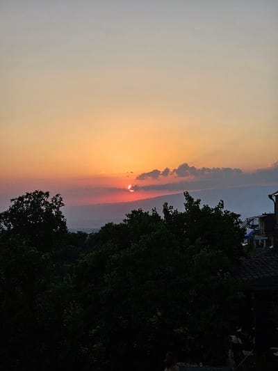 This image captures a sunset view within the city of Dzoraghbyur, Armenia. The upper portion of the frame is dominated by a clear sky displaying a gradient of colors, transitioning from light blue-grey at the top to vibrant orange, red, and pink hues near the horizon. The sun is partially visible as a bright, reddish-orange disc, mostly obscured by a band of dark, irregular clouds positioned along the horizon line. Faint crepuscular rays extend upwards from the sun's position. Below the horizon, a distant, undulating dark silhouette suggests mountains or hills, partially obscured by atmospheric haze. The lower half of the image is dominated by a dense canopy of dark green trees, heavily silhouetted against the brighter sky, obscuring the ground plane. On the far right, a portion of a building is visible, including a dark, shingled roof in the lower right corner and a lighter-colored structure above it, which appears to feature a balcony or upper level and a blue slide-like object. No individuals or specific activities are discernible.